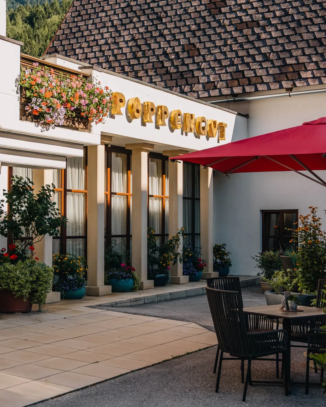 © Hartwig Winkler Der Eingang des Poppengut im Sommer, der goldene Schriftzug über dem Vordach, eine Blumenkiste, ein roter Sonnenschirm und ein Tisch auf der Terrasse