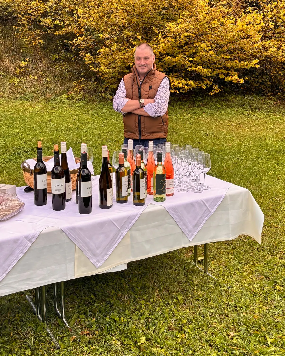 © Hartwig Winkler Ein Mann in brauner Weste steht hinter einem weiß gedeckten Tisch mit Weinflaschen und Gläsern auf einer Wiese, dahinter gelbes Herbstlaub