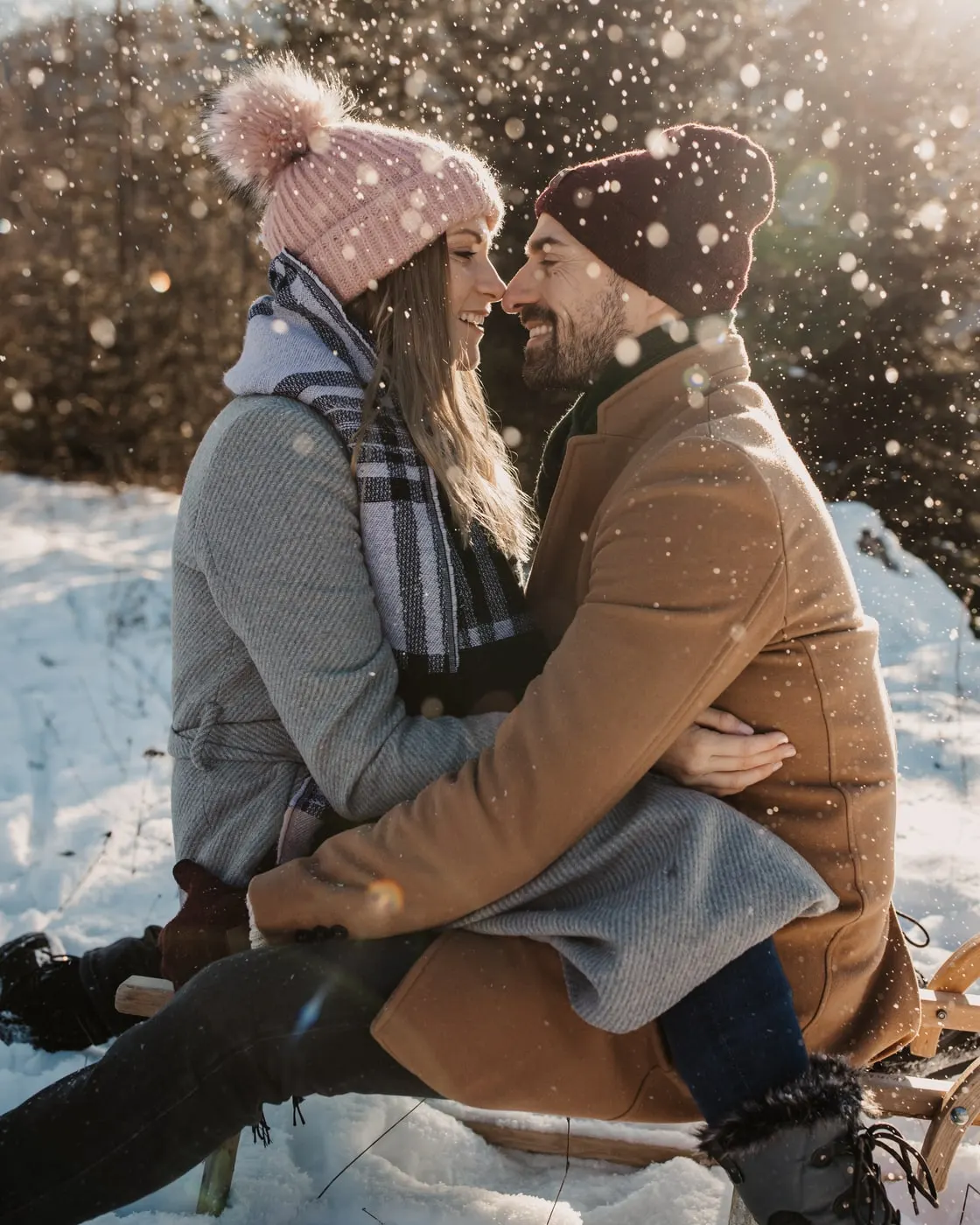 © Selina Flasch Photography Lachendes Paar mit Wintermützen Nase an Nase vor einem Nadelwald, ringsum glitzern Schneeflocken in der Sonne