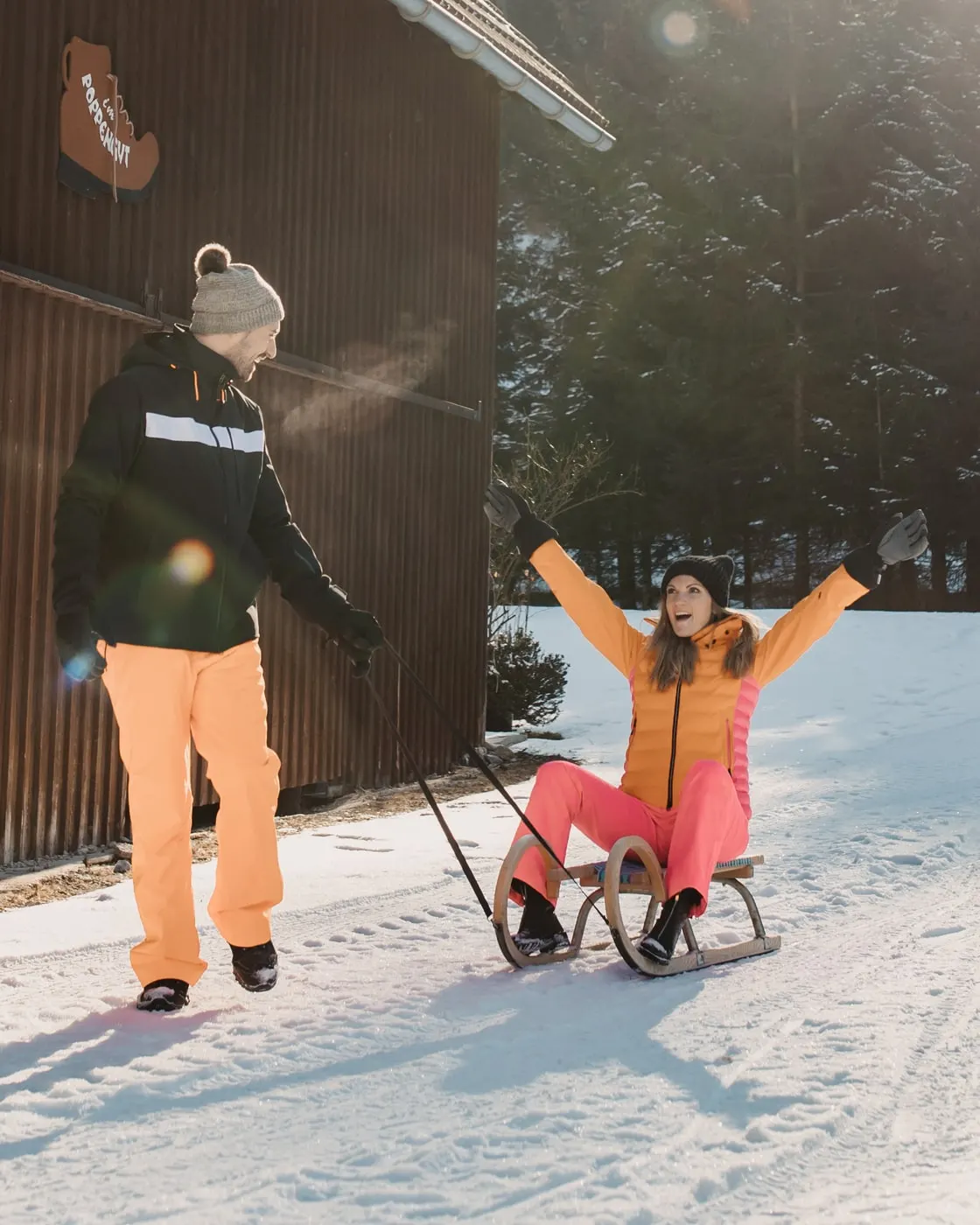 © Selina Flasch Photography Ein Paar beim Rodeln vor einer Holzscheune im Schnee: Er zieht die Rodel, sie sitzt darauf und jubelt mit erhobenen Armen
