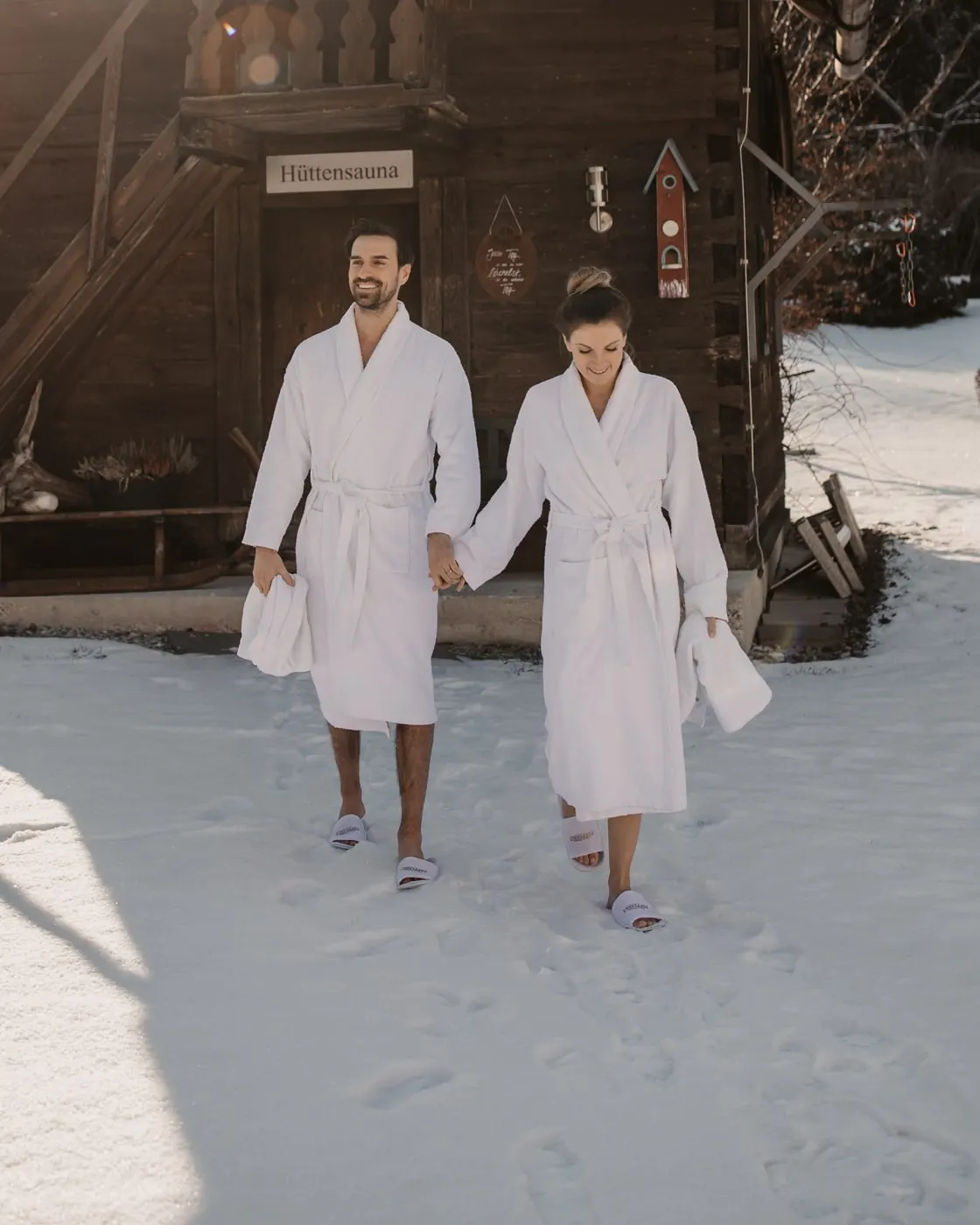 © Selina Flasch Photography Zwei Gäste in weißen Bademänteln gehen Hand in Hand durch den Schnee, hinter ihnen die hölzerne Hüttensauna
