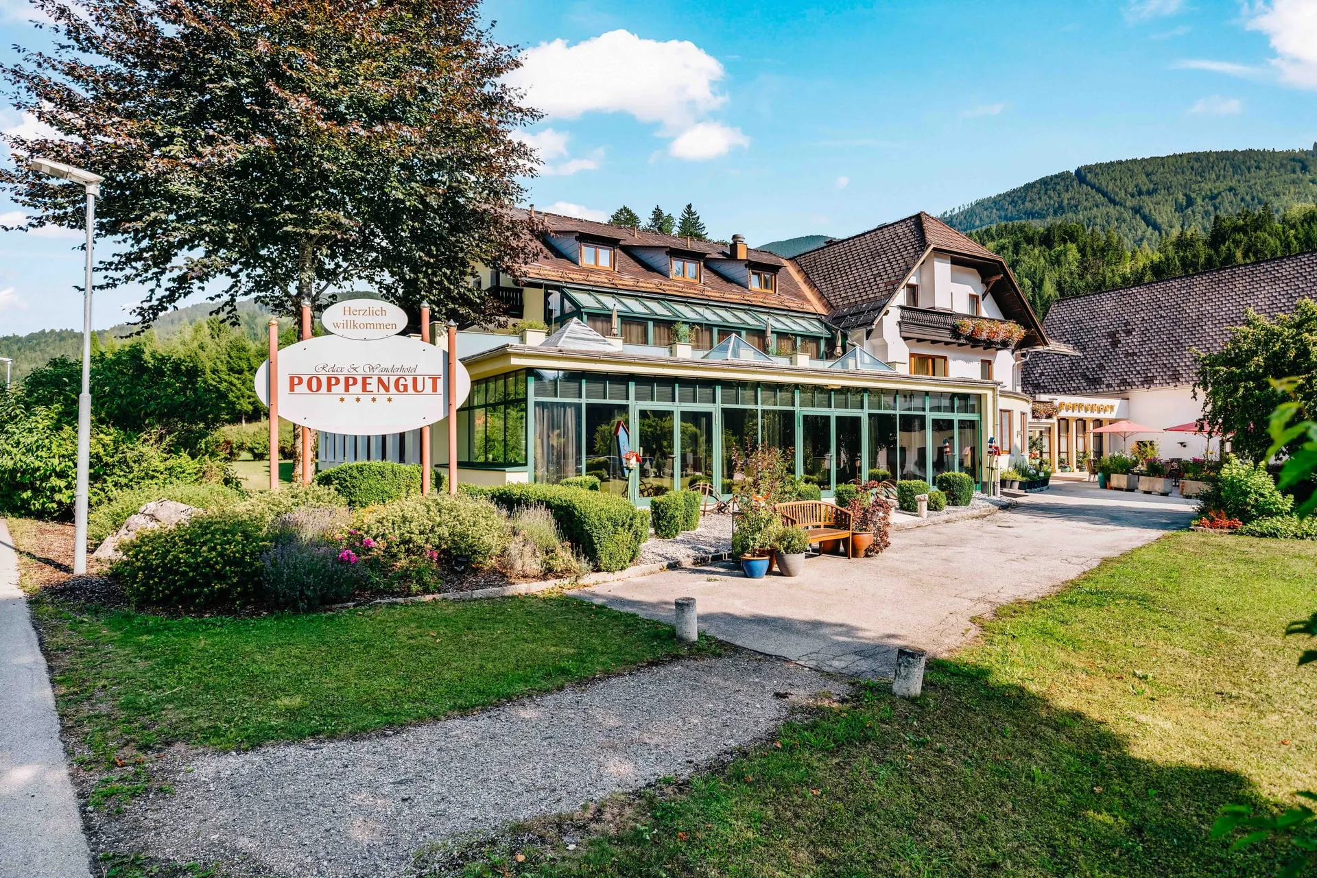 © Hartwig Winkler Der Eingang des Poppengut im Sommer – das Schild „Herzlich willkommen“, der Wintergarten und dahinter das Haus